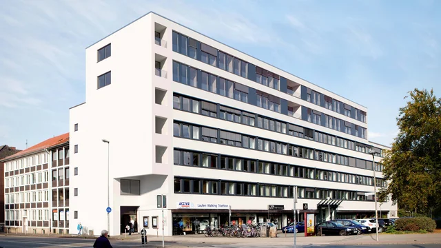 Αρχιτεκτονικός φωτορεαλισμός του σύγχρονου κτιρίου κατοικιών και καταστημάτων στη Blissestraße 5 στο Βερολίνο, με λευκή πρόσοψη, μεγάλες τζαμαρίες και αστικό περιβάλλον δρόμου.